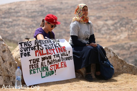 פעילה יהודיה אמריקאית ואשה פלסטינית יושבים יחד. על השלט שהן אוחזות כתוב "היהודיה האמריקאית הזאת תומכת בצדק לפלסטין. שברו את החומה, שחררו את כל האסירים הפוליטיים . "צומוד, מחנה החירות". הכפר סארורה הגדה המערבית, 19 במאי 2017 (צילום: אחמד אל-באז אקטיבסטילס)